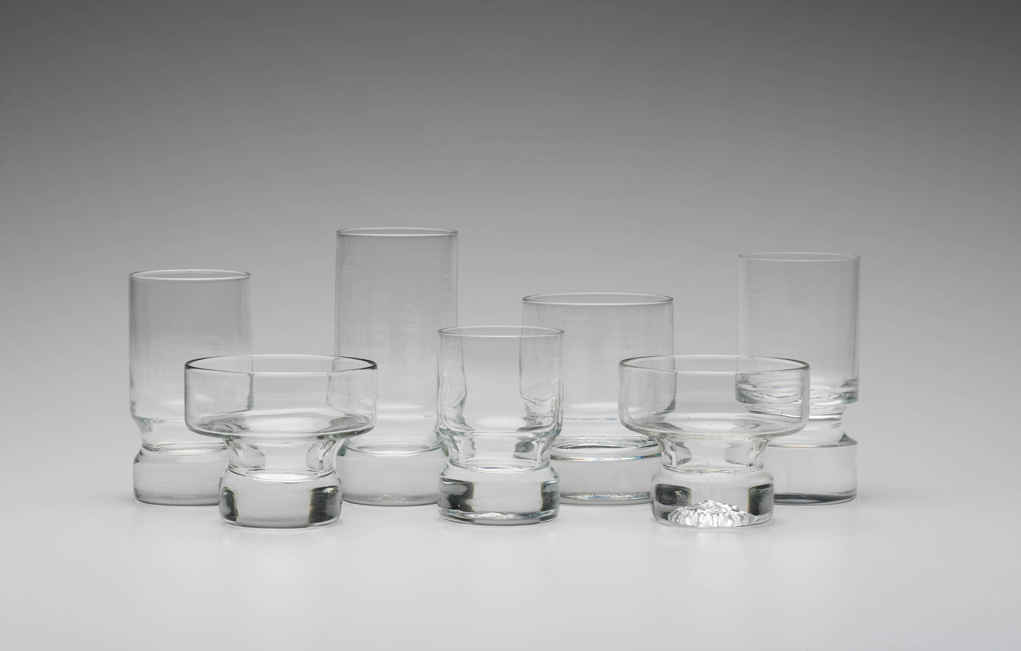 Seven drinking glasses of different shapes and sizes on a grey background.
